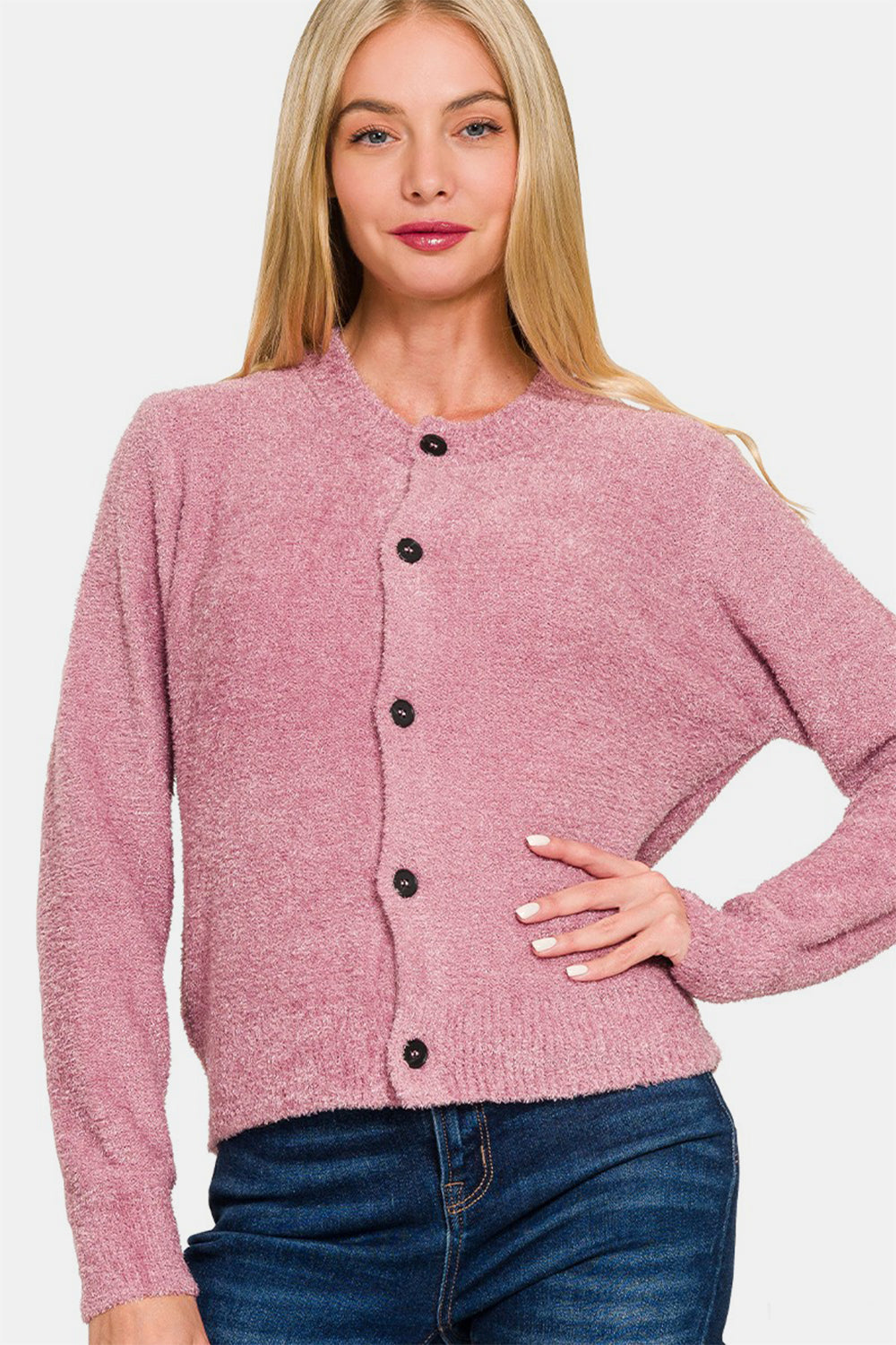 A person with long blonde hair is wearing a Zenana Button Down Long Sleeve Sweater Cardigan in pink over a white crop top and blue jeans, posing with one hand touching their hair.