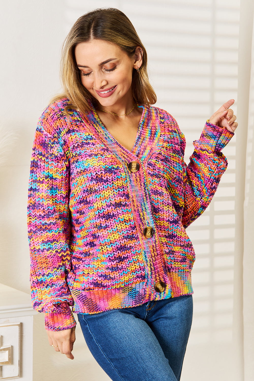 Person wearing an Angel Wings Woven Right V-Neck Long Sleeve Cardigan, characterized by its multicolored design and large buttons, paired with blue jeans. The cardigan, made of 100% acrylic knit, offers a versatile look perfect for any occasion. They are standing indoors against a white background.