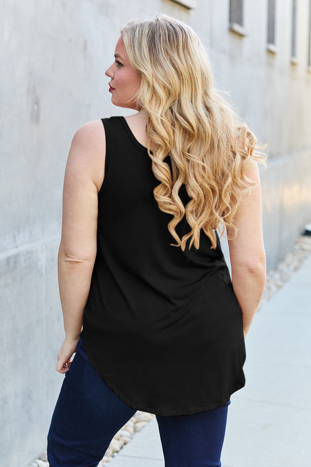 A woman wearing sunglasses, blue jeans, and the Basic Bae Full Size Round Neck Curved Hem Tank is standing in front of a concrete wall, holding her glasses with one hand and carrying a beige shoulder bag. Her basic style complements the slightly stretchy fabric of her outfit, ensuring comfort while maintaining chic simplicity.