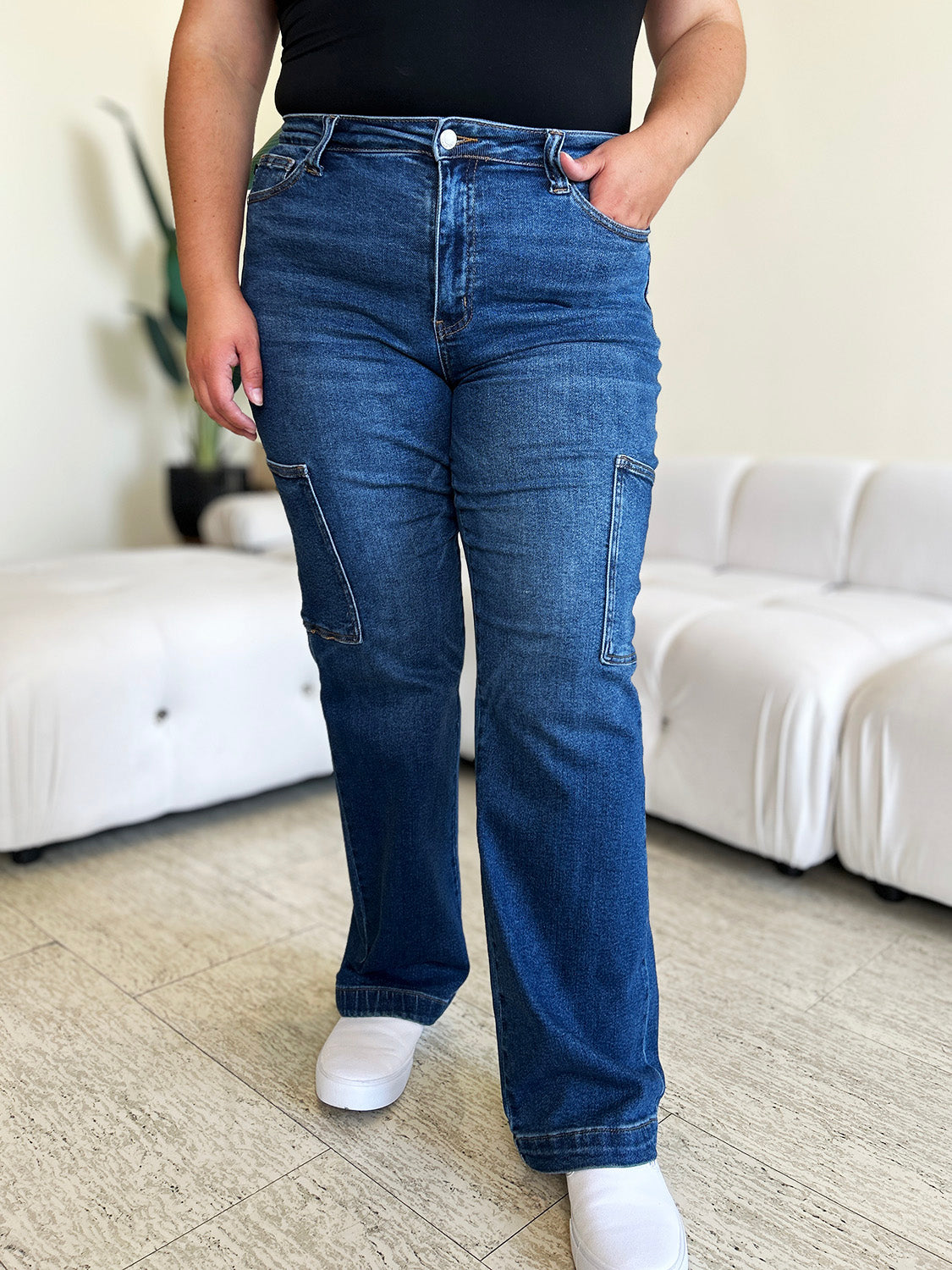 A person wearing Judy Blue Full Size High Waist Straight Cargo Jeans made of durable denim fabric and a white top walks on a wooden floor, with a white sofa and green plant in the background.