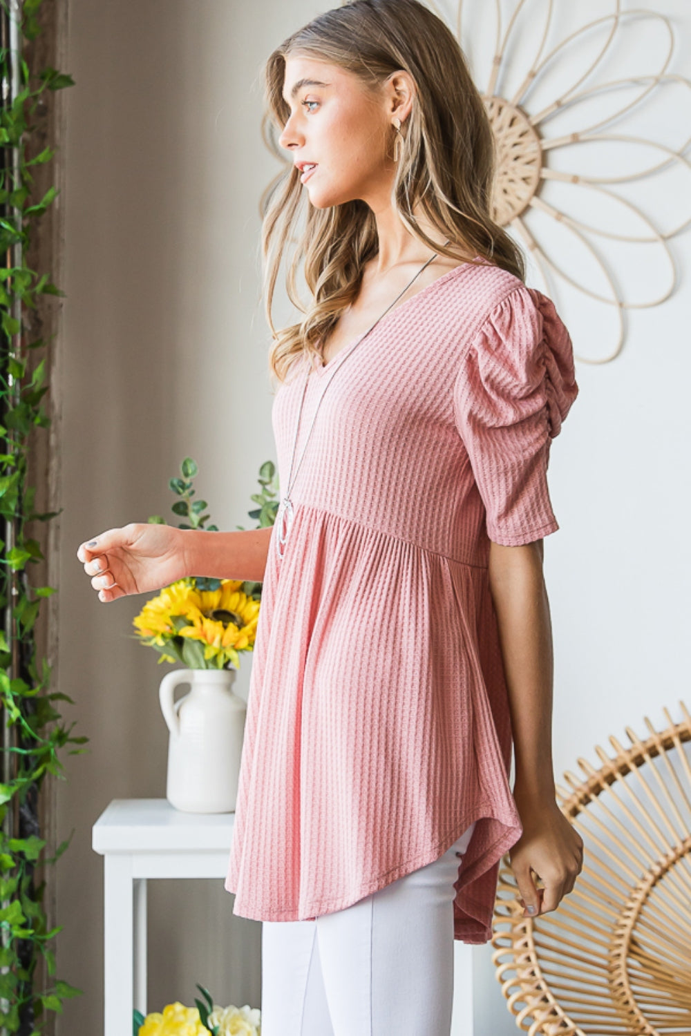 A woman in a Heimish Full Size Waffle Knit V-Neck Babydoll Top with short-sleeves and a long necklace, standing by a window with a neutral expression; the perfect cozy wardrobe addition.