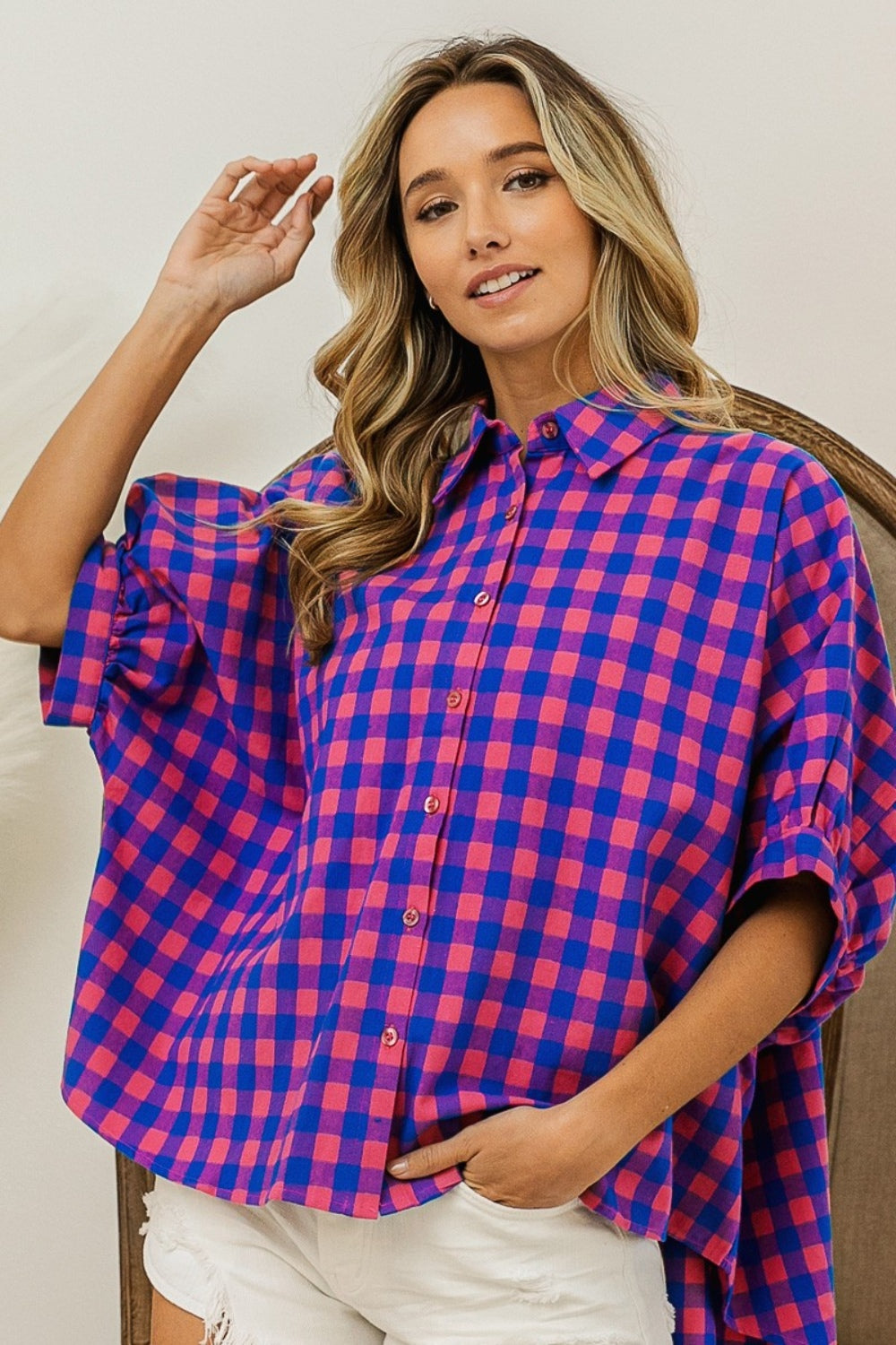 Person wearing a BiBi Plaid Button Up Dolman Sleeve Shirt in blue and pink with white shorts, standing in front of a wooden chair.