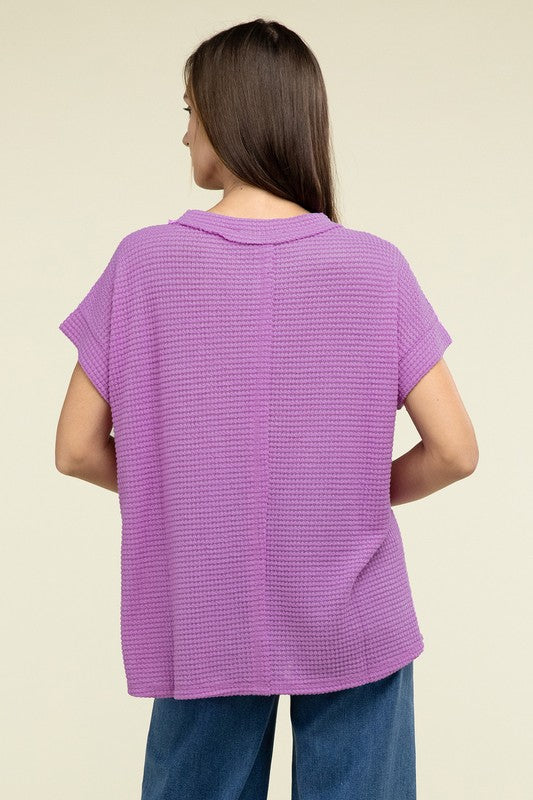 A woman with long brown hair is wearing a purple Brushed Waffle Exposed-Seam Short Sleeve Top and blue jeans, standing against a plain beige background. This casual wardrobe choice, with its short sleeves and side slits, is perfect for everyday wear.