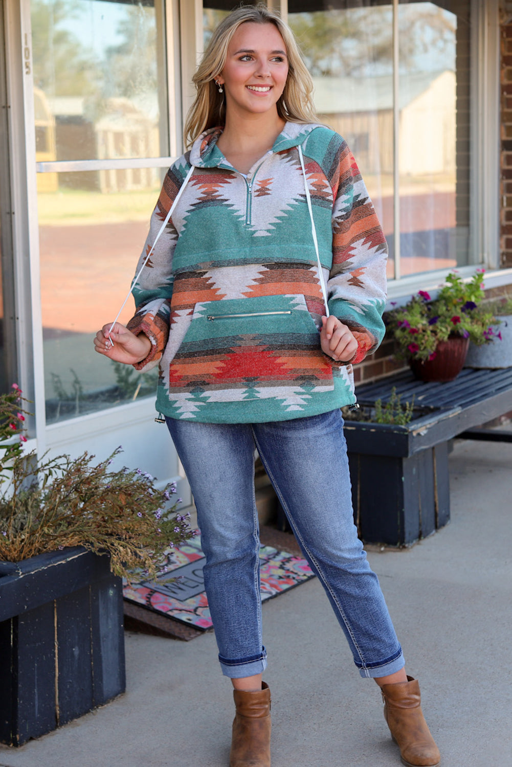 A woman with long blonde hair is standing outside, facing away from the camera. She wears a Multicolor Aztec Print Zipped Split Neck Hoodie and jeans. A rug and potted plant are visible in the background, adding to the cozy vibe of her super comfortable outfit.