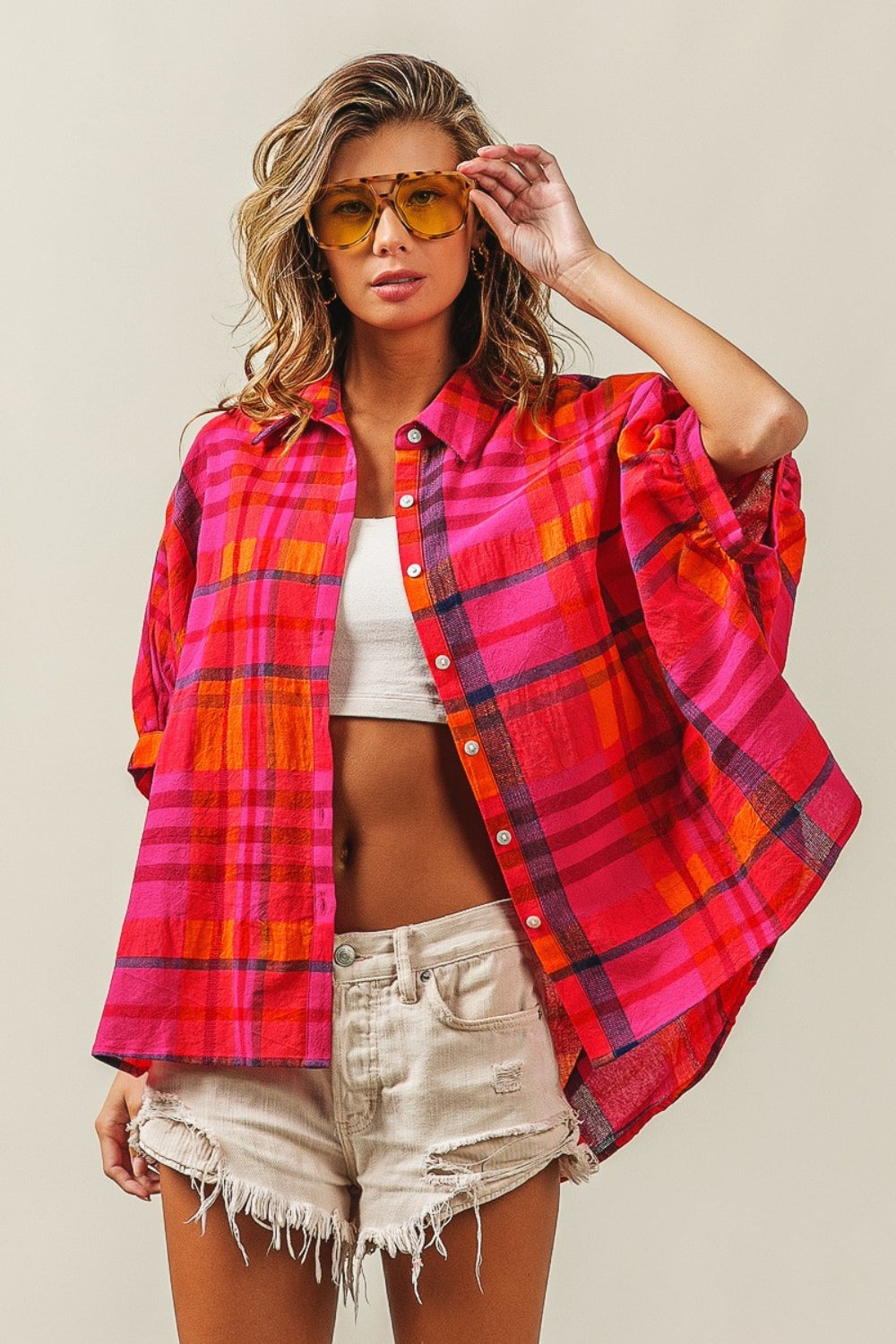 A woman outfitted in a BiBi Button Up Dolman Sleeve Plaid Shirt, paired with a white crop top, distressed denim shorts, and yellow sunglasses.