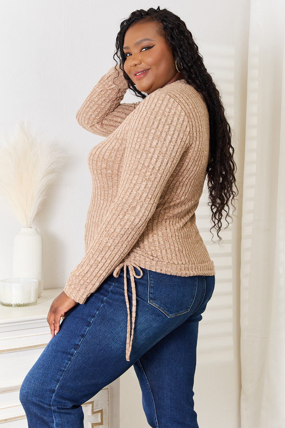 A woman stands indoors against a neutral background, wearing the "Double Take Drawstring Ribbed Long Sleeve T-Shirt" in light purple paired with dark pants. She is smiling and posing with one hand on her hip.