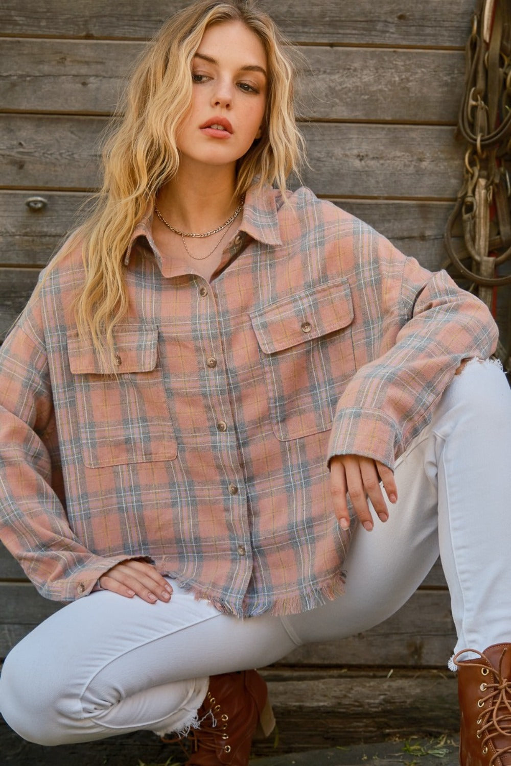 A person wearing the And The Why Full Size Plaid Button Up Raw Hem Shirt and white pants is standing in front of a wooden structure, showcasing raw edge detailing, with bridles hanging beside them.