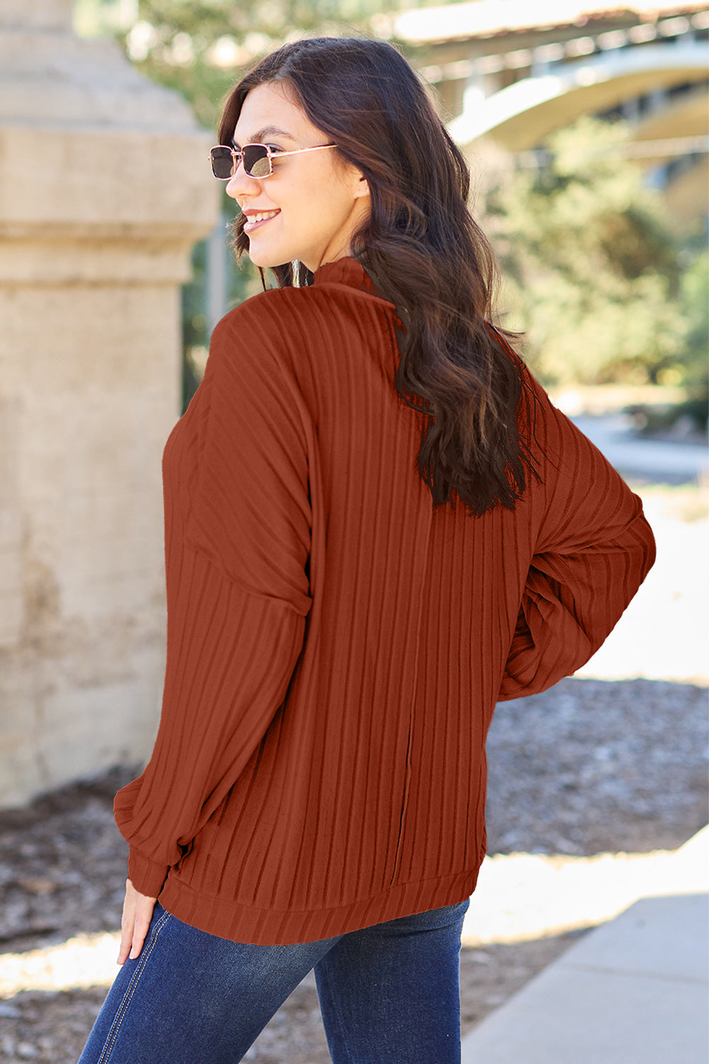 A woman with long hair is wearing a green Basic Bae Full Size Ribbed Exposed Seam Mock Neck Knit Top, blue jeans, and sunglasses. She is standing outdoors and smiling.