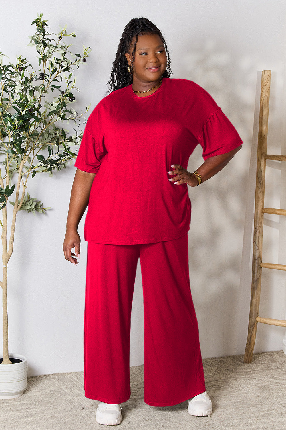 A woman stands near a potted plant and wooden ladder, wearing the Double Take Full Size Round Neck Slit Top and Pants Set in a bright pink two-piece outfit with matching wide-leg pants that are available in plus size and are easily machine washable.