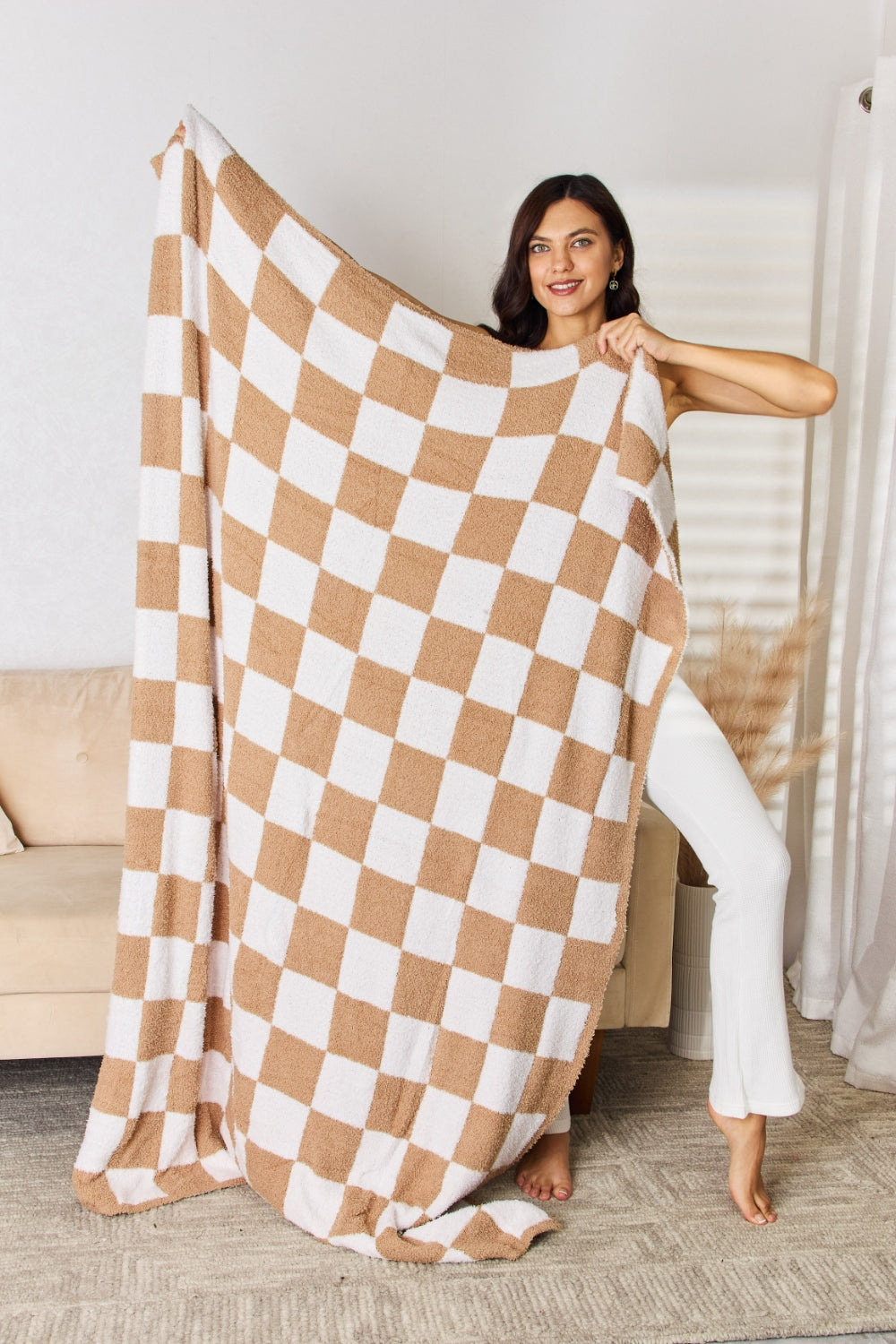 A person stands holding a large Cuddley Checkered Decorative Throw Blanket, smiling, in a room with a sofa and a potted plant. The blanket features a black and white checkerboard pattern, is made from 100% polyester, and imported for quality.