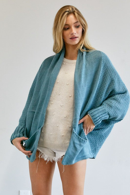 A person stands against a plain background, wearing a Pattern Knit Dolman Sleeve Solid Slouch Cardigan in textured blue over a white sweater with distressed white shorts, hands in cardigan pockets.