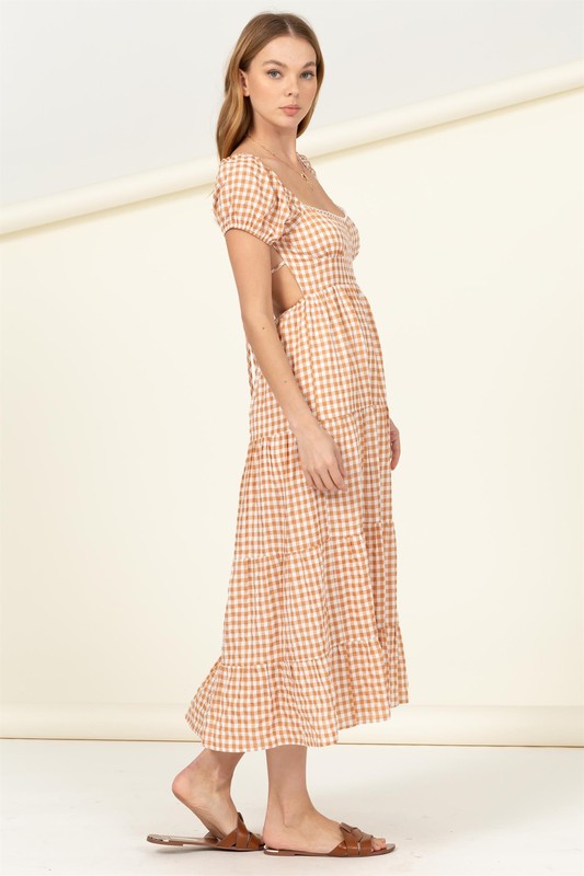 Wearing the "Somewhere to Go Tie-Back Gingham Print Maxi Dress," which features a black and white gingham print with puffed sleeves, a tiered skirt, and elegant tie-back detail, a person poses stylishly paired with black sandals.