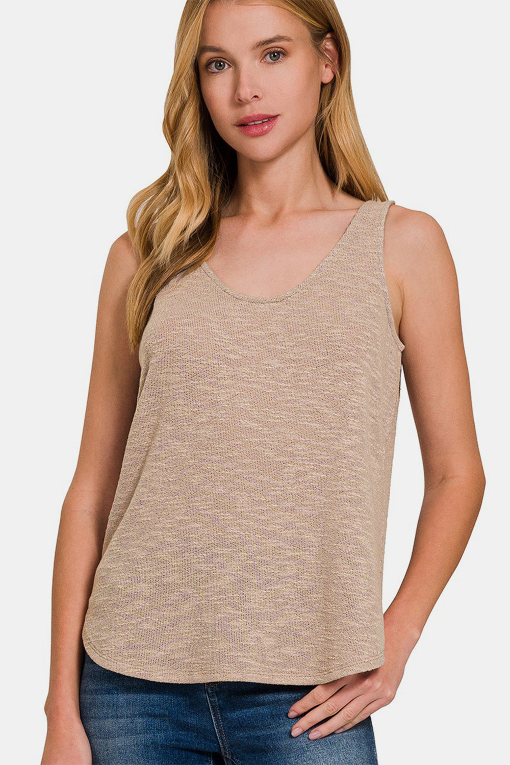 A woman with long, blonde hair wears a Zenana Curved Hem Round Neck Tank in beige and blue jeans, perfect for a casual wardrobe, standing against a plain background.