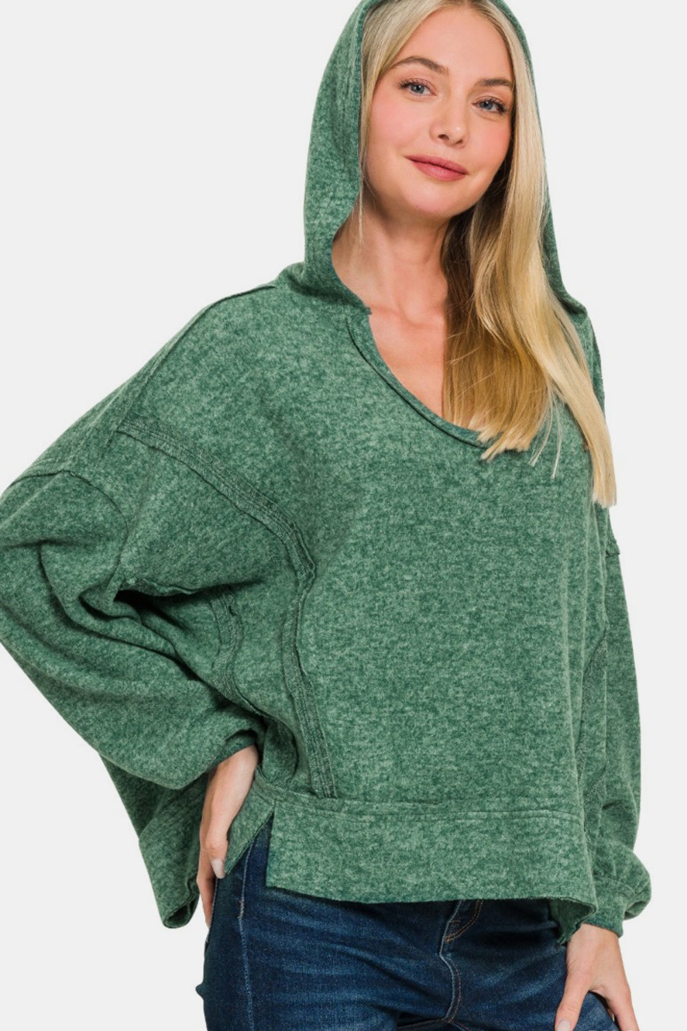 A person wearing a Zenana Brushed Hacci Exposed Seam Hoodie in green, complemented by jeans, poses confidently with a hand on their hip.