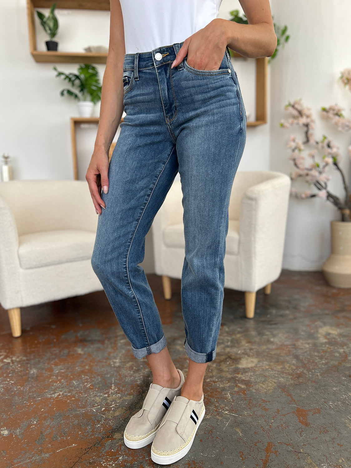 A person wearing a white shirt, Judy Blue Full Size Cuffed Hem Slim Jeans, and black wedge-heeled sandals is standing in a room with beige chairs and plants in the background.