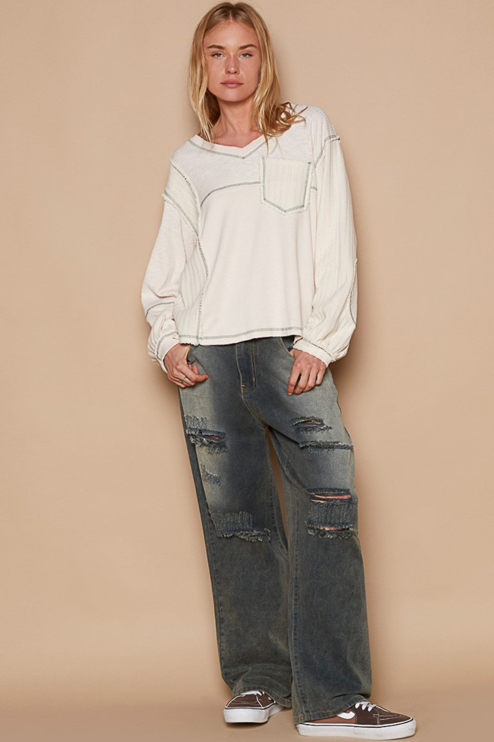 A person wearing the POL V-Neck Knit Panel Exposed Seam Top in cream and distressed jeans stands against a tan background.