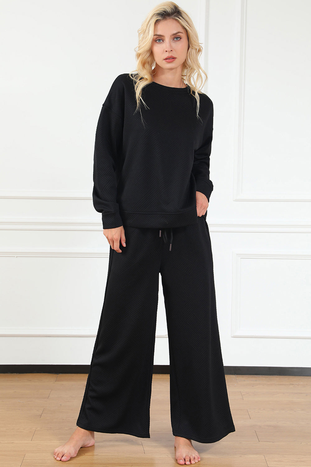 A woman with blonde hair, dressed in the Double Take Full Size Textured Long Sleeve Top and Drawstring Pants Set featuring a stretchy black long-sleeve top and matching wide-leg trousers made of rayon spandex, stands barefoot in a room with white walls and a wooden floor.