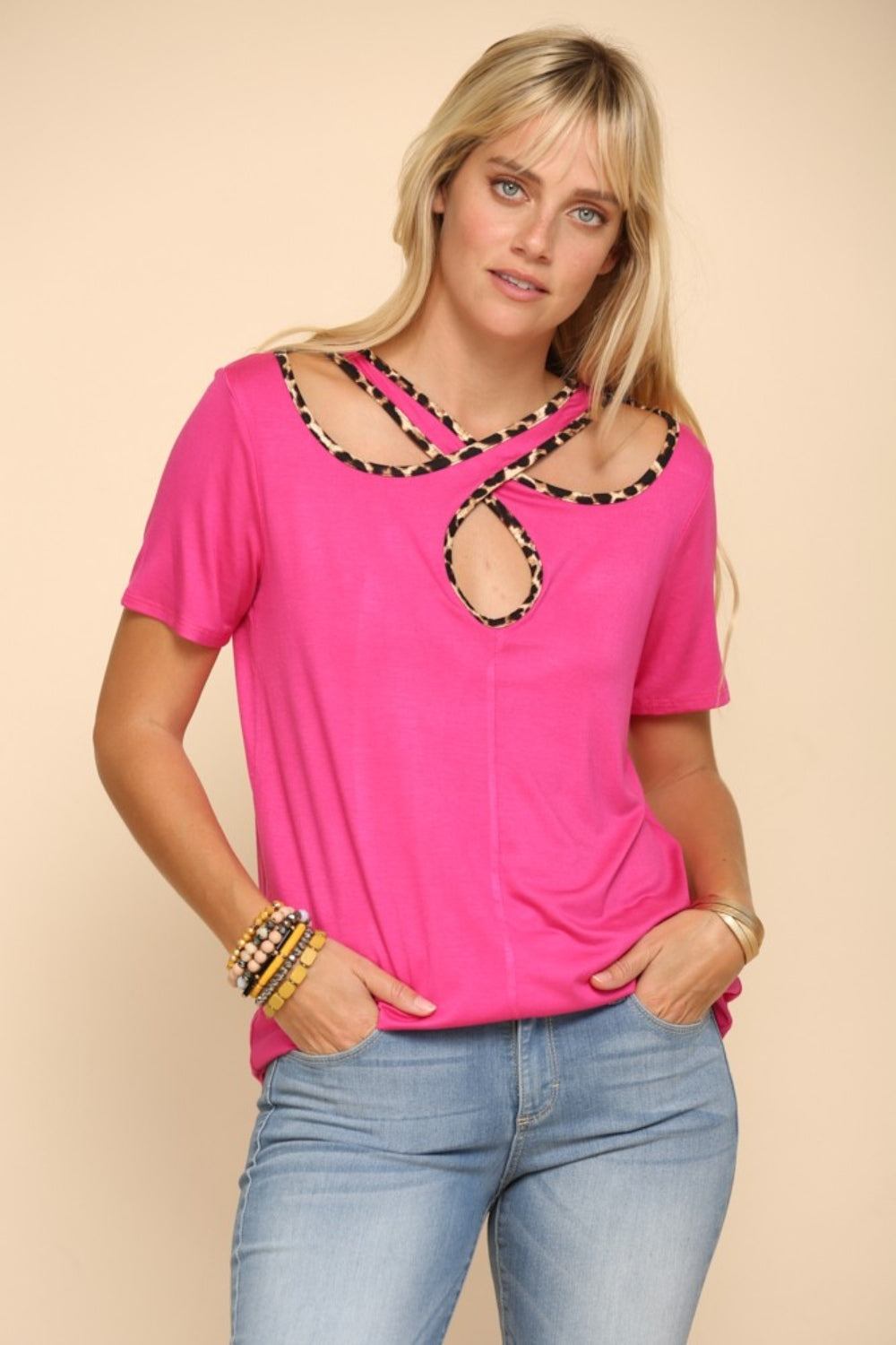 A person showcases the Celeste Full Size Crisscross Cutout Leopard Contrast Trim T-Shirt in bright pink, paired with light blue jeans, posing confidently against a beige background.