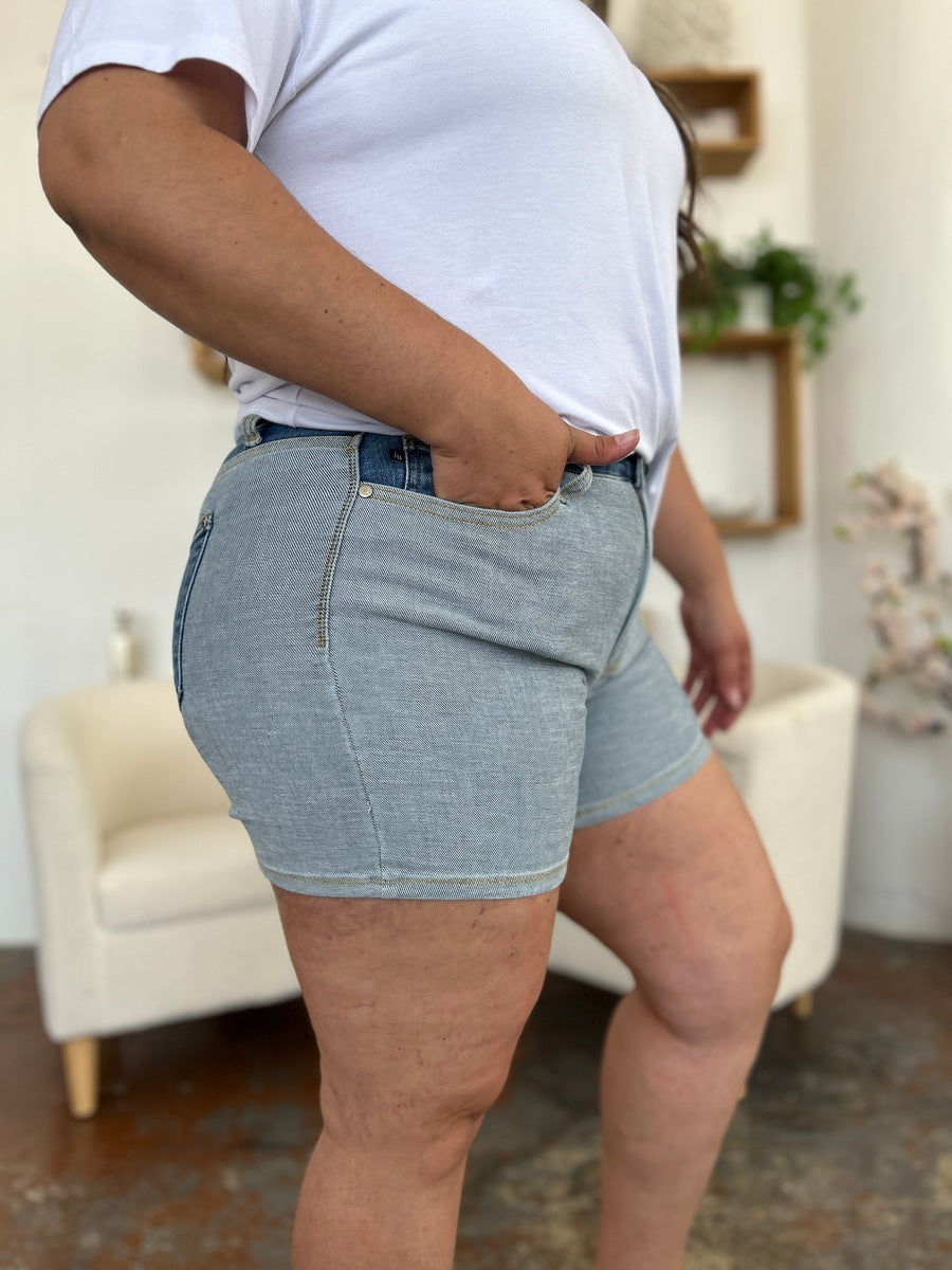 A person models indoors beside a beige chair and a plant, sporting a white t-shirt with the Judy Blue Full Size Color Block Denim Shorts for an effortlessly bold, eye-catching look.