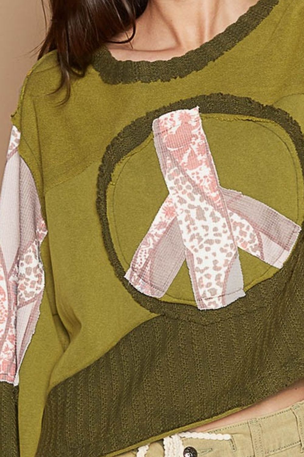 A woman with long hair is standing against a beige background, wearing the POL Long Sleeve Cut Sew Peace Emblem Top in olive green and beige pants.