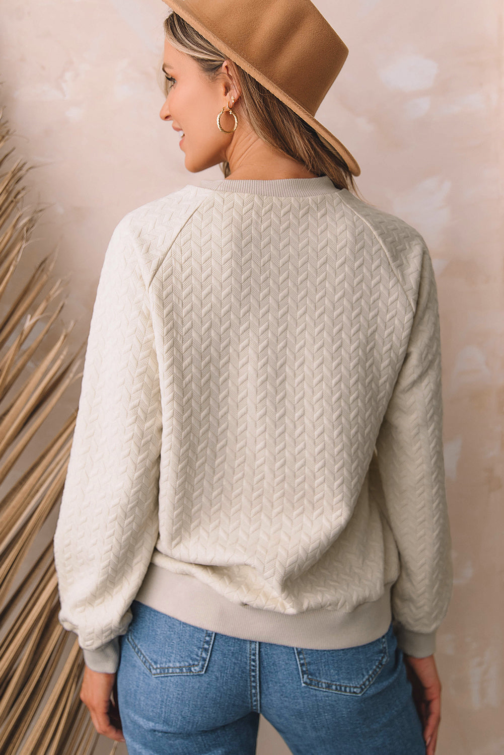 A woman in a Beige Solid Textured Raglan Sleeve Pullover Sweatshirt and blue jeans is looking to the side. She sports a brown hat and earrings, exuding a casual and comfy vibe. Standing next to dried pampas grass against a light background, she seamlessly blends with her textured sweatshirt.