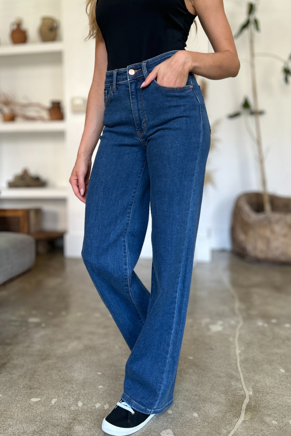 A person wearing Judy Blue Full Size High Rise Straight Jeans, a black top, and white shoes stands gracefully on a polished concrete floor.