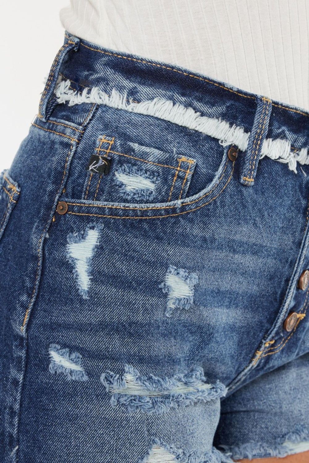 A woman with curly hair wearing a white sleeveless top and Kancan Distressed Button Fly Denim Shorts stands against a plain background, perfect for a stylish summer wardrobe.