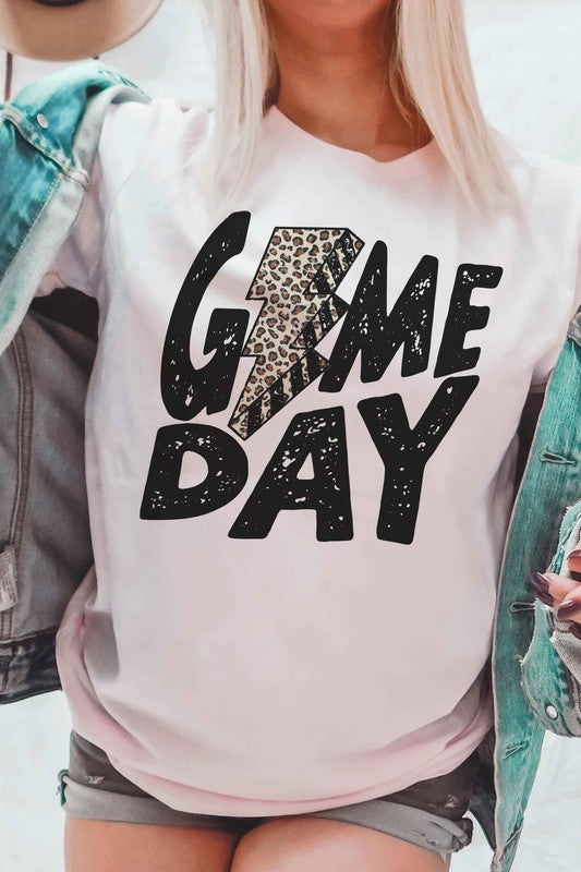 A person wearing a white "LEOPARD LIGHTNING GAME DAY Graphic Tee" with a leopard print lightning bolt design stands with a denim jacket partially on, showcasing its unisex sizing.