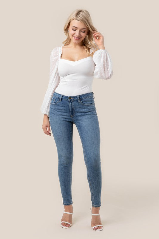 A person wearing the LS shirring sleeve bodysuit with Swiss dot mesh, featuring sheer, long puffed sleeves and a sweetheart neck, paired with light-colored jeans, stands against a plain background.