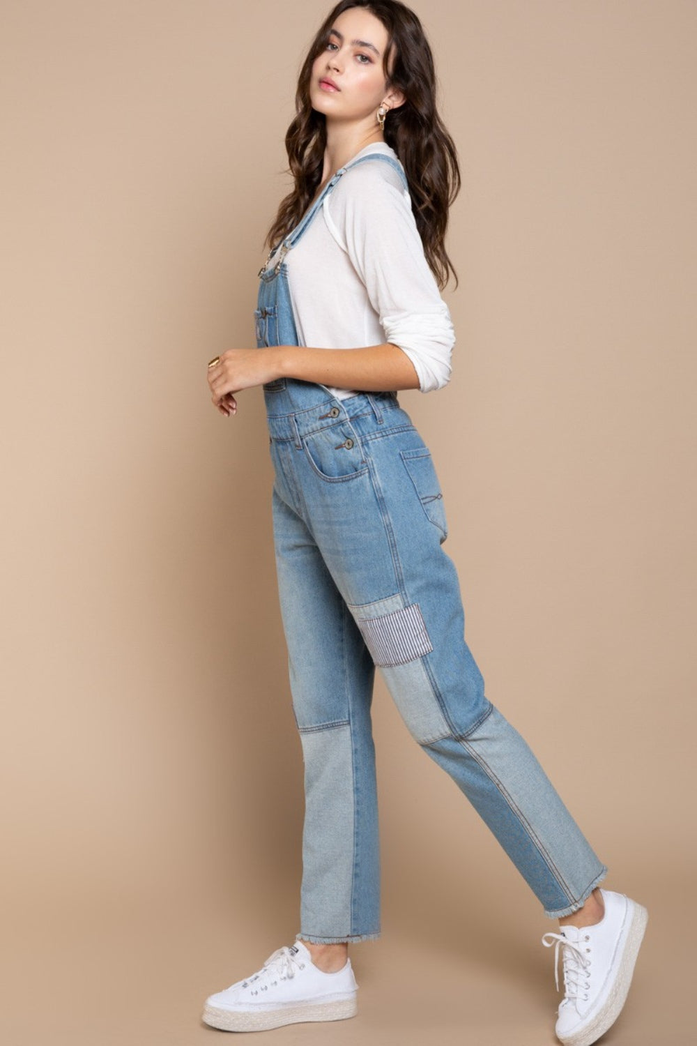 A woman kneels on one knee against a beige background, wearing a beige hat and white long-sleeve shirt, paired with POL Front Chest Zipper Slim Leg Denim Overalls featuring patches.