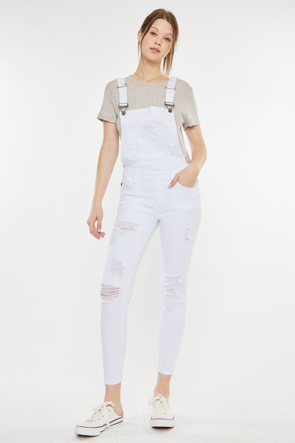 A fashion-forward woman stands against a plain background, wearing a grey t-shirt, Kancan Distressed Skinny Denim Overalls in white, and white sneakers with red detailing.