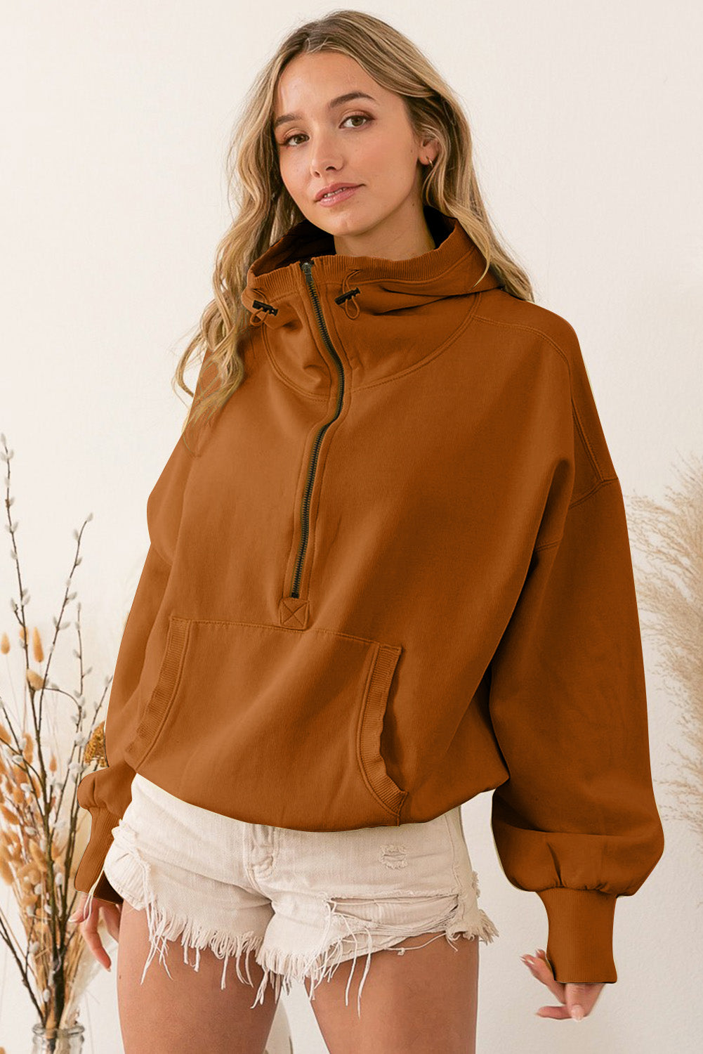 A person wearing a Brown Ribbed Trim Kangaroo Pocket Zipped Hoodie and frayed shorts stands sideways near decorative dried plants in vases.