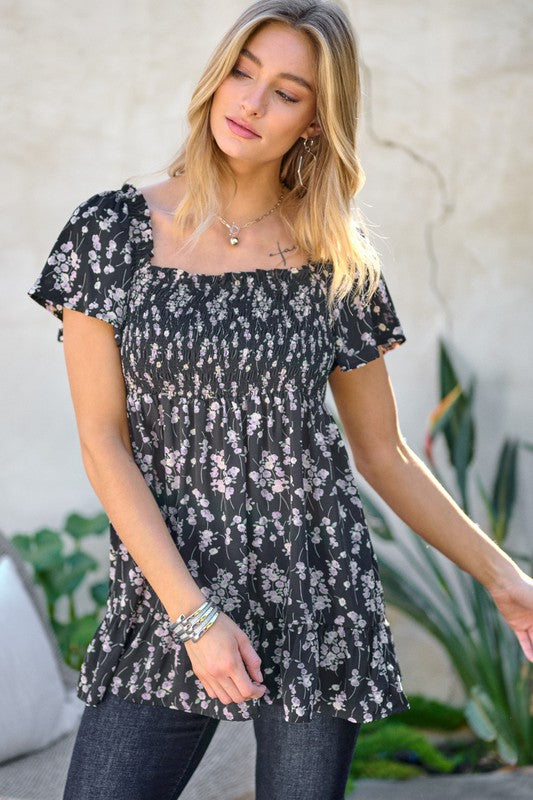 A woman stands outdoors donning a Floral Printed V-Neck Ruffle Top paired with jeans, set against a neutral background, with plants in the foreground, highlighting her fashion-forward wardrobe.