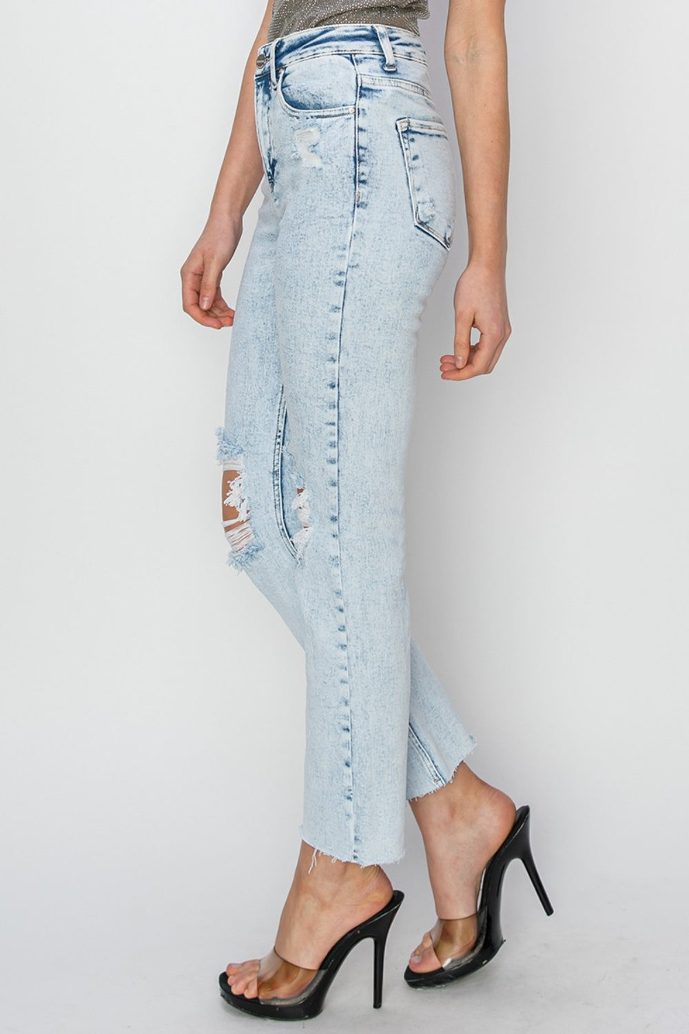 Person wearing RISEN High Rise Distressed Ankle Jeans in light blue with rips and raw hems, paired with transparent-strap high heels.