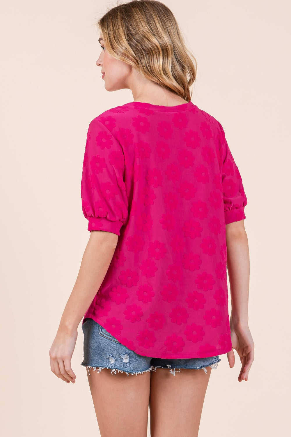 A woman showcases her versatile style in the BOMBOM Textured Floral Pattern Top, with its bright pink hue and feminine design, paired effortlessly with denim shorts against a plain background.