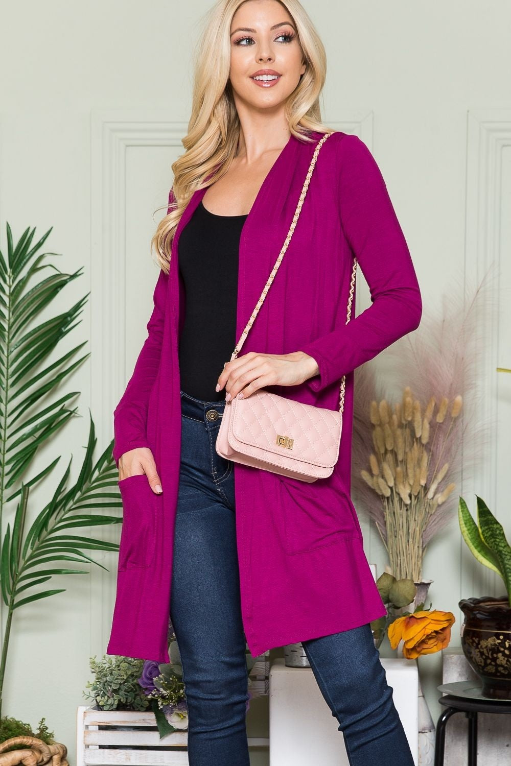 A woman with long blonde hair, wearing the Celeste Full Size Open Front Cardigan with Pockets in pink and a black top, poses with a light pink quilted purse on her shoulder. The open-front design of her versatile layering piece complements the indoor plants and light green wall behind her perfectly.