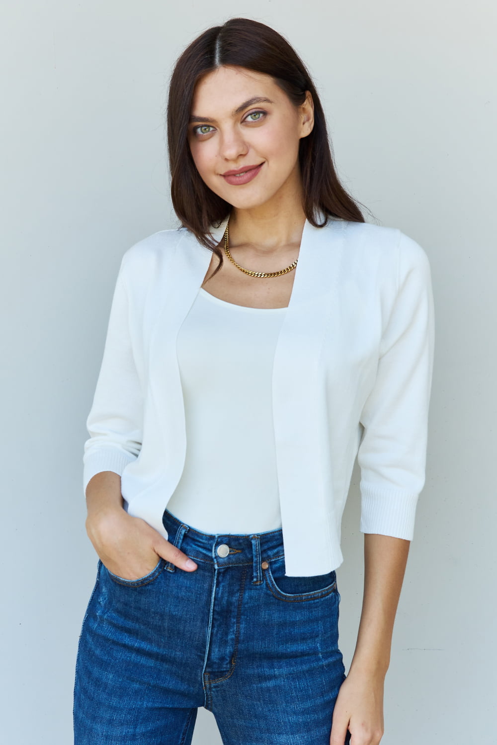 A stylish person wearing sunglasses, a Doublju My Favorite Full Size 3/4 Sleeve Cropped Cardigan in Ivory, and blue jeans poses in front of a plain background, effortlessly highlighting their versatile look.