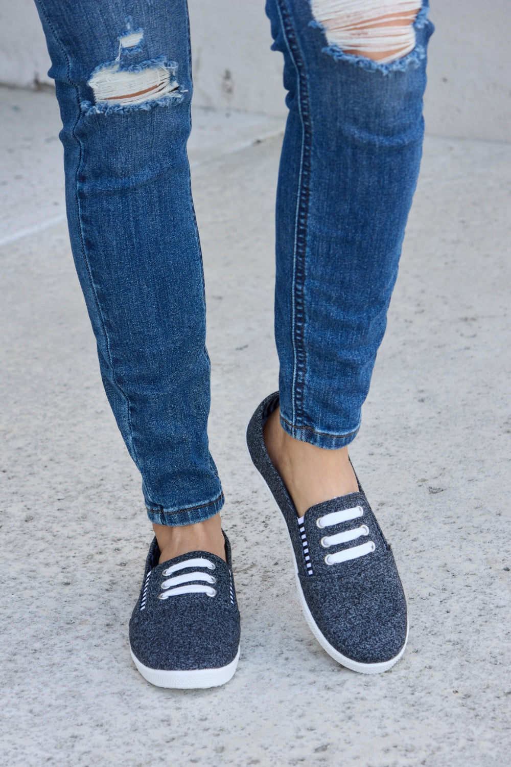 A person wearing ripped blue jeans and the fashionable Forever Link Round Toe Slip-On Flat Sneakers with white elastic bands.