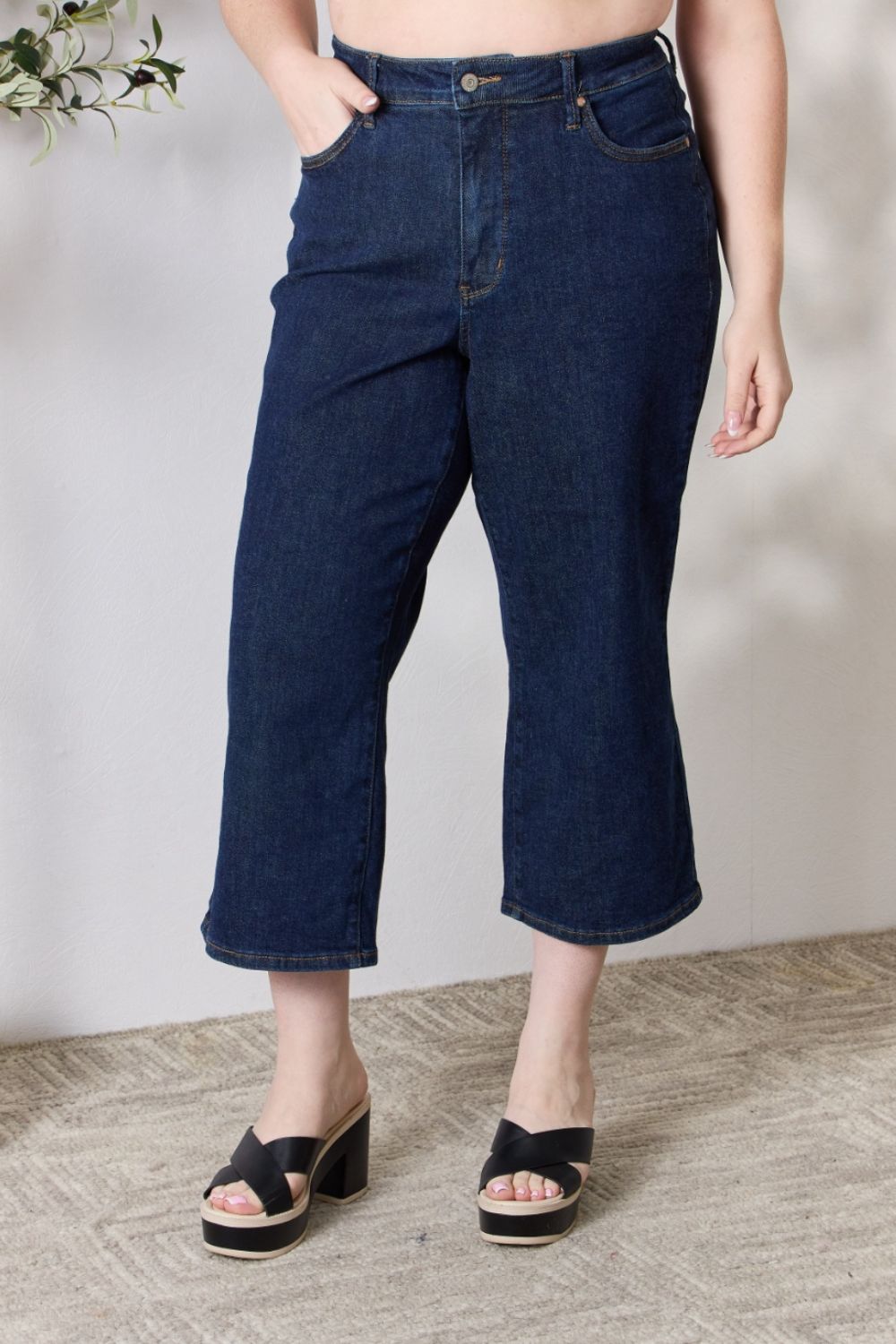 A person wearing the Judy Blue Full Size High Waist Cropped Wide Leg Jeans, which are renowned for their tummy control design, and nude block heel sandals stands near a potted plant.