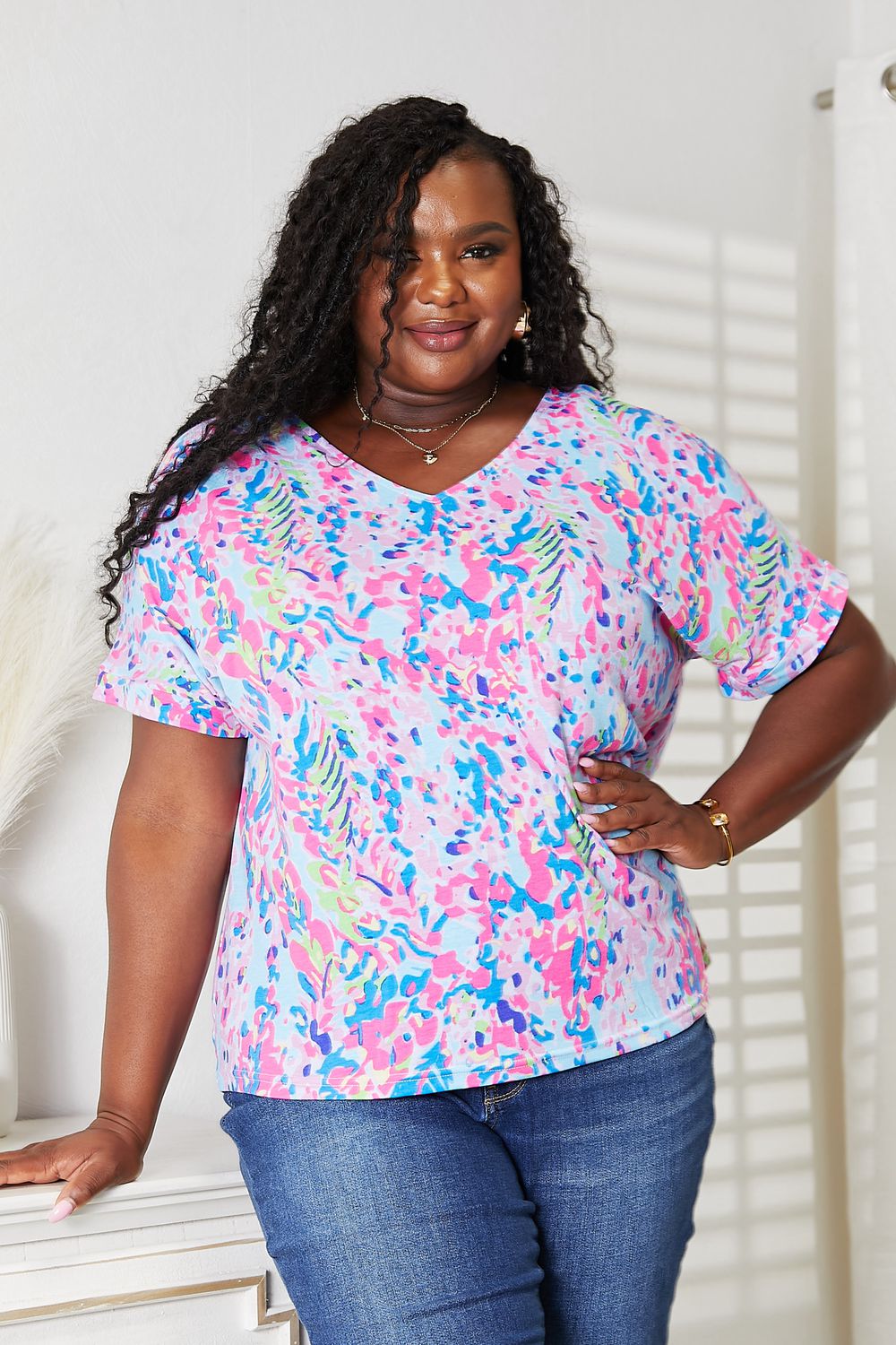 A person with long hair smiles while wearing a vibrant Printed V-Neck T-Shirt and jeans. They stand in a well-lit room with a white wall and a decorative plant, highlighting the colorful design of their top.