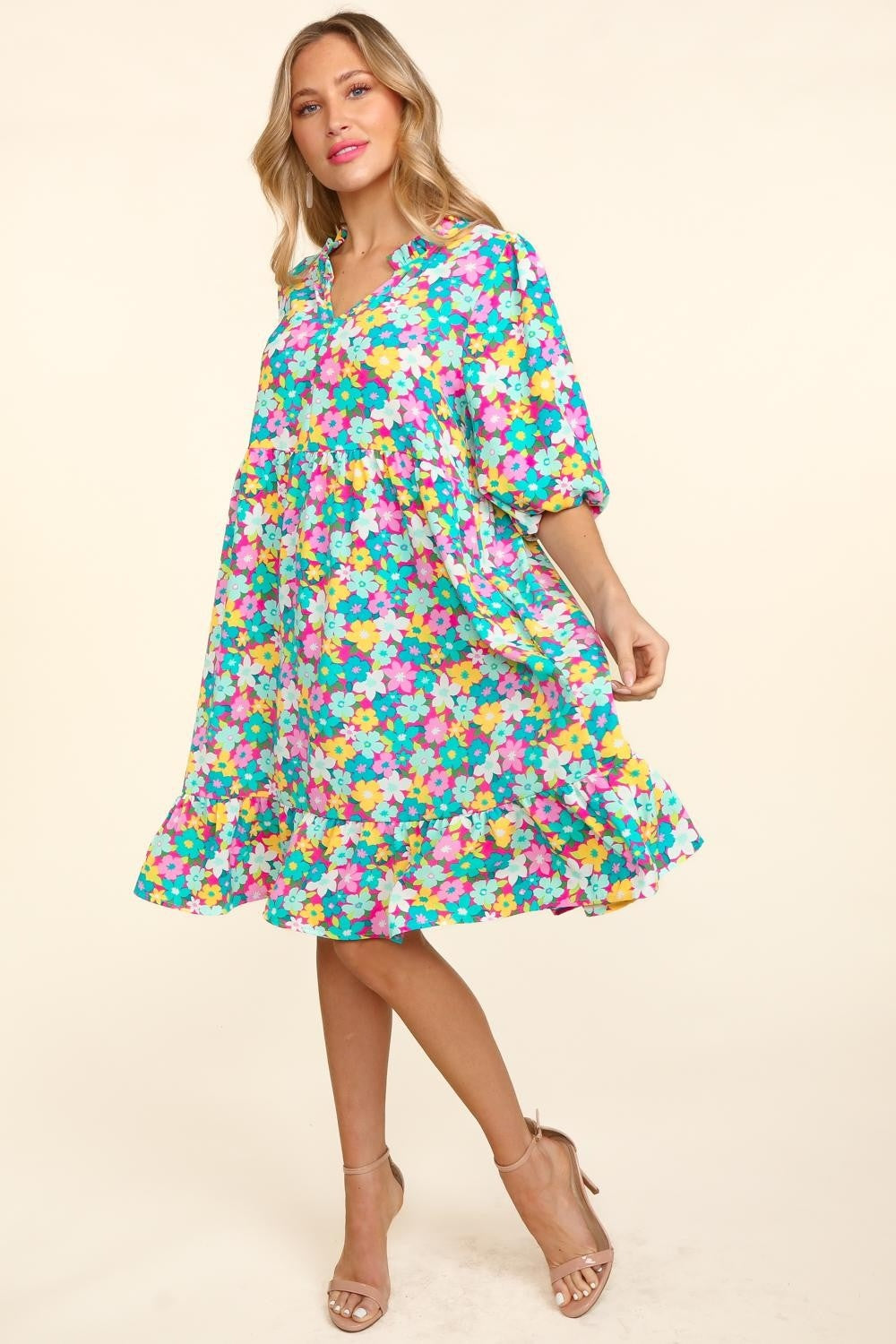 A woman in a Haptics Bubble Sleeve Floral Ruffled Dress stands against a plain background, with one hand holding her dress and the other by her side. She has long, wavy hair and is wearing open-toed shoes, perfect for any garden party.