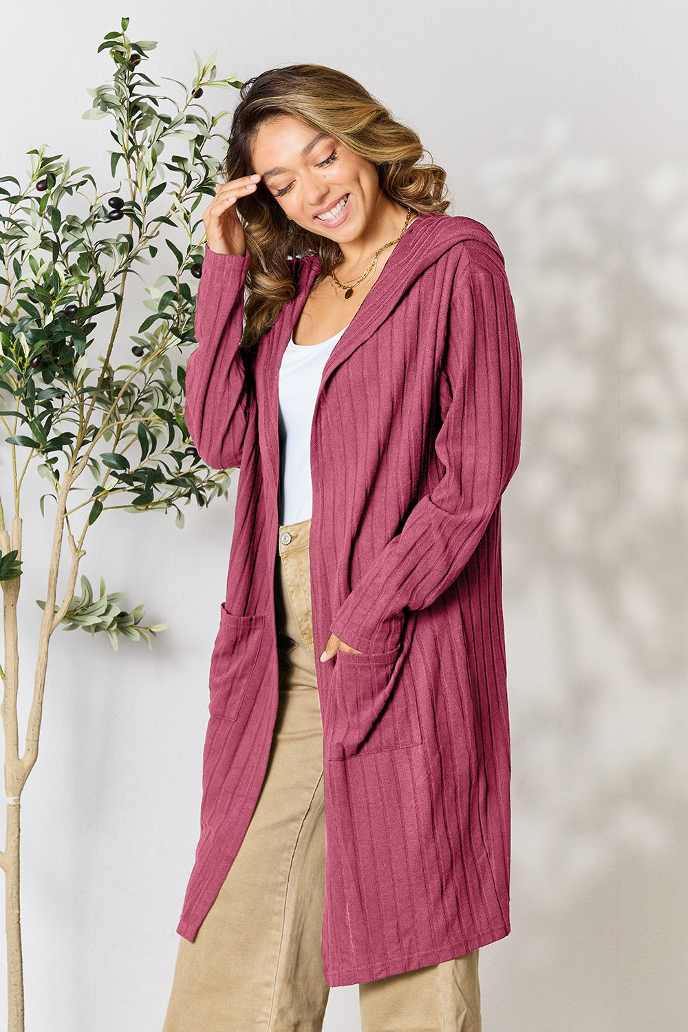 A woman wearing the Basic Bae Full Size Hooded Sweater Cardigan in black, paired with a white top and khaki pants, stands in front of a light background accented by a leafy plant on her left. With one hand tucked into her pocket, she gazes to her right.