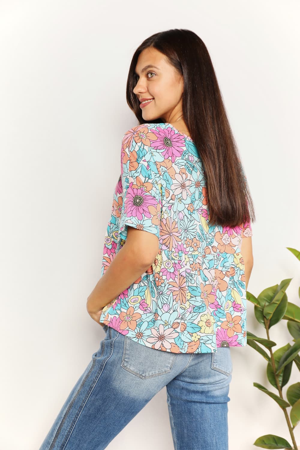 A person wearing a Double Take Floral Round Neck Babydoll Top and jeans stands with one hand in their pocket, with a plant in the background.