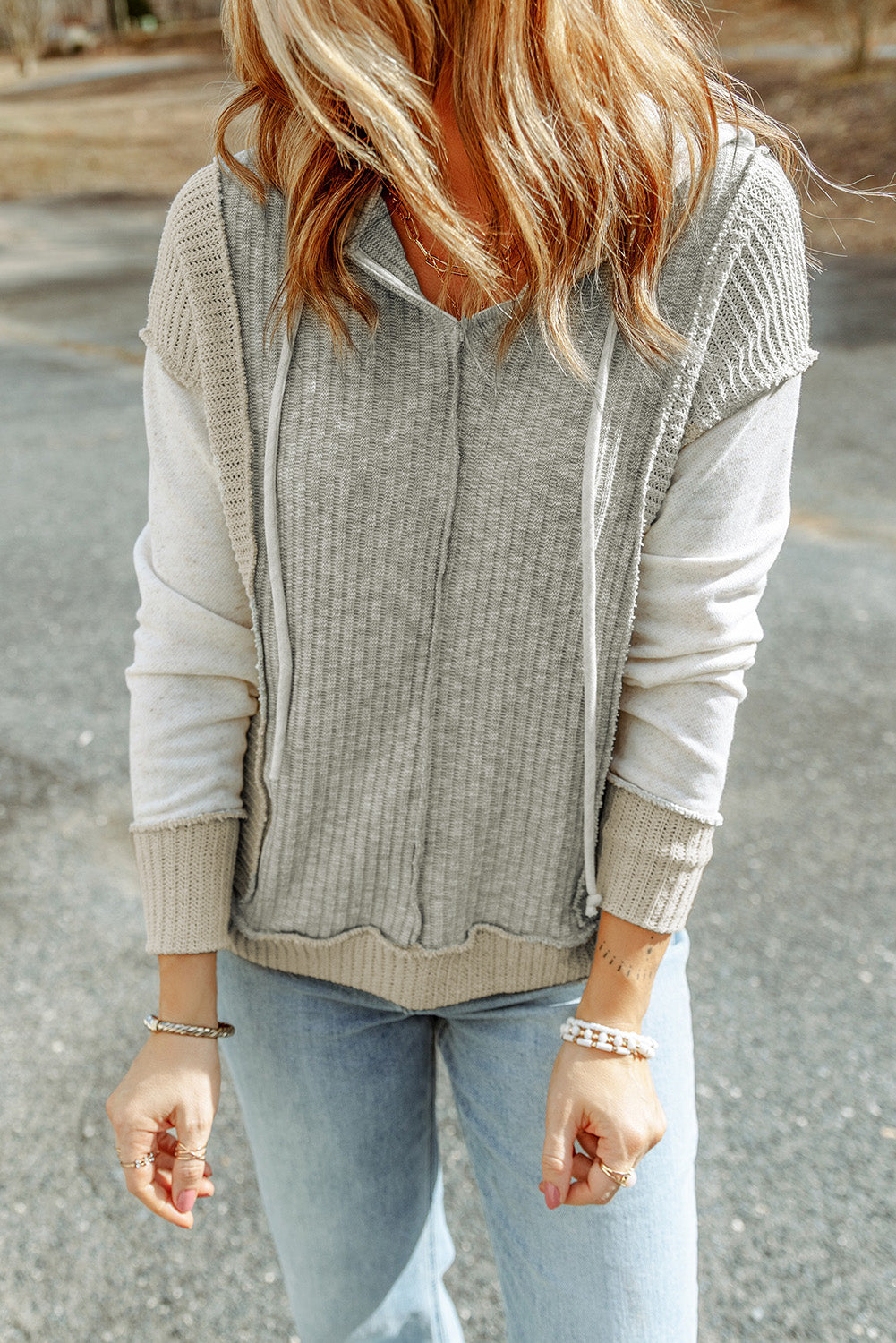 Back view of a person with long, wavy hair wearing a Gray Long Sleeve Textured Knit Patchwork Hoodie and light blue jeans, standing on a paved surface. The person is holding a strand of hair with one hand and has the other hand on their hip.