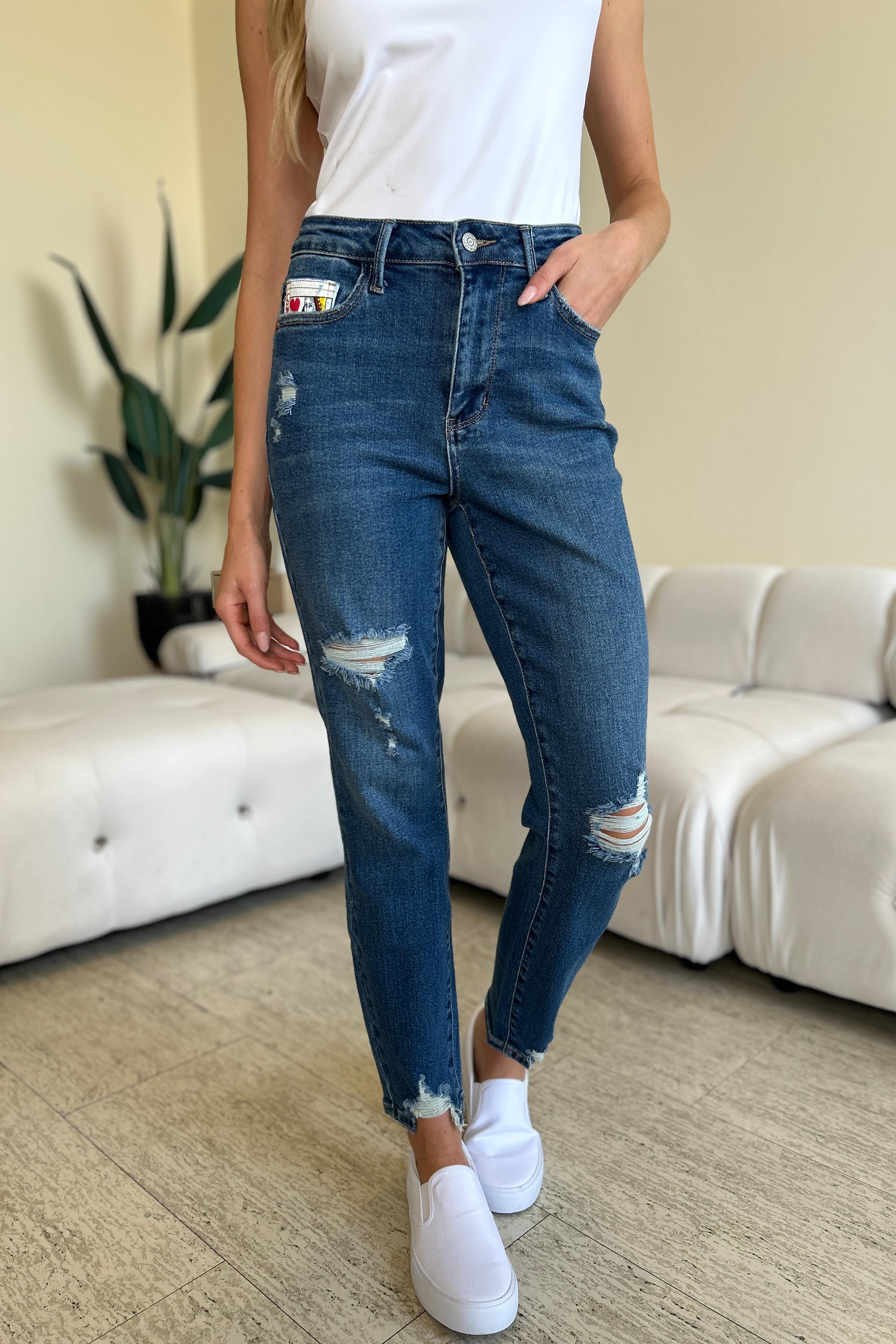 Two women stand side by side in a living room, both wearing stylish jeans and tank tops. One woman is pointing at the distinctive back pocket of her Judy Blue Full Size Queen Of Hearts Coin Pocket BF Jeans, while the other smiles.