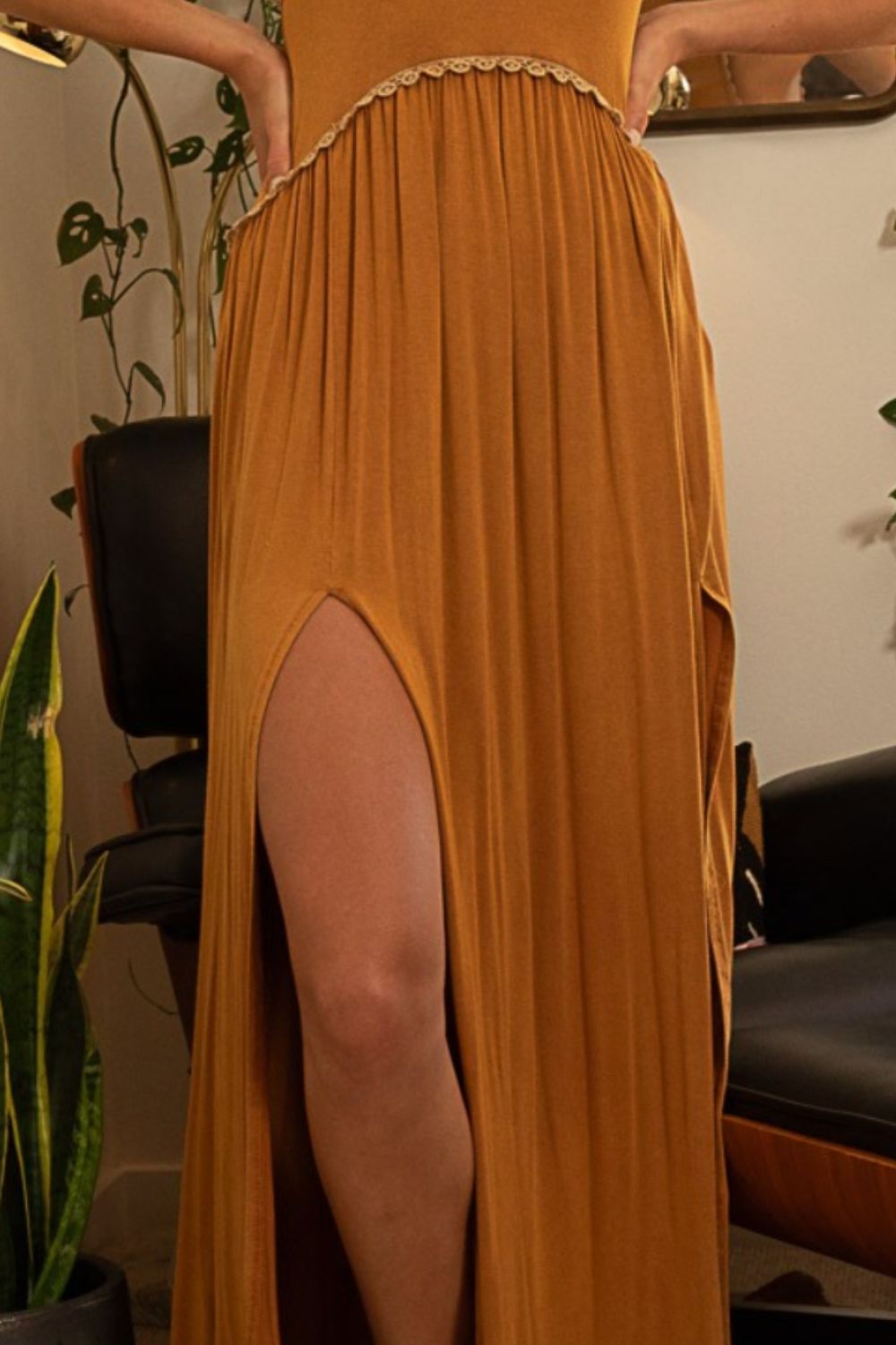 A woman stands barefoot in a room wearing a POL Sleeveless Back Zipper Front Slit Maxi Dress in mustard yellow, leaning against a doorframe. The versatile dress adds to her wardrobe, complemented by plants and a chair in the background.