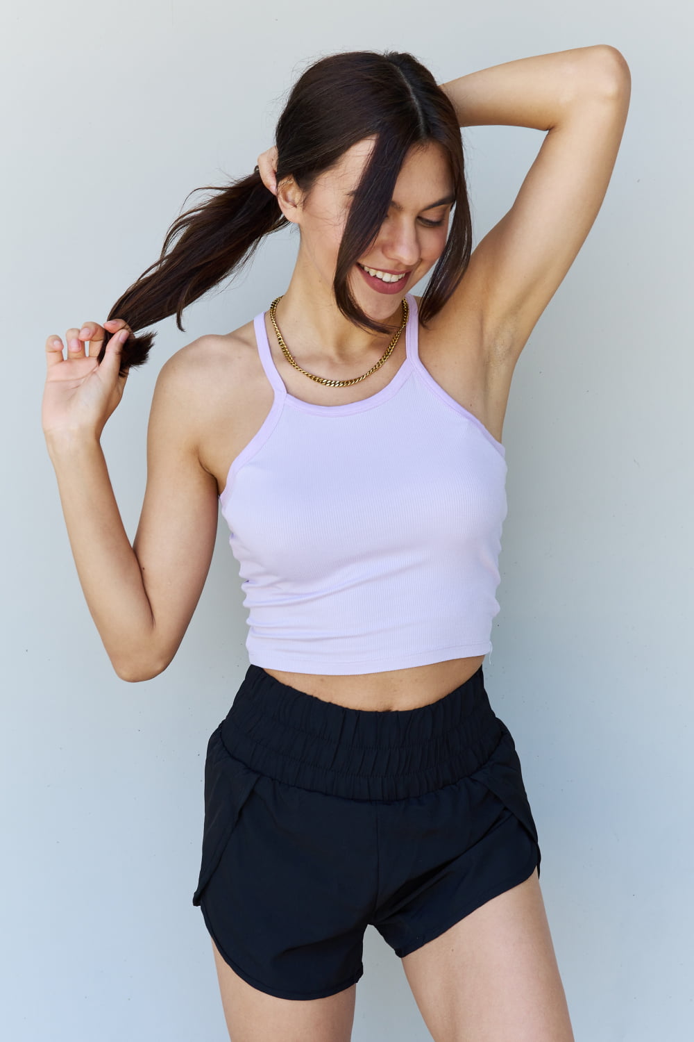 A woman with long brown hair, wearing the Ninexis Everyday Staple Soft Modal Short Strap Ribbed Tank Top in Lavender and black shorts, poses while smiling in front of a plain background.