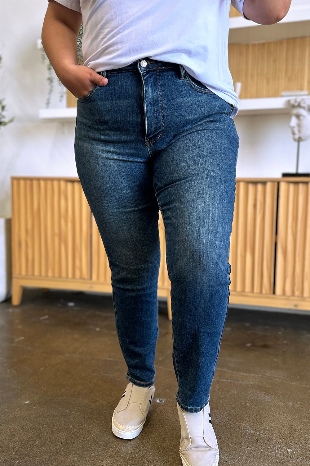 Person wearing Judy Blue Full Size Tummy Control High Waist Slim Jeans in blue and a white shirt, with their hands in their pockets. They are standing indoors on a polished concrete floor with a wooden cabinet and plant in the background.