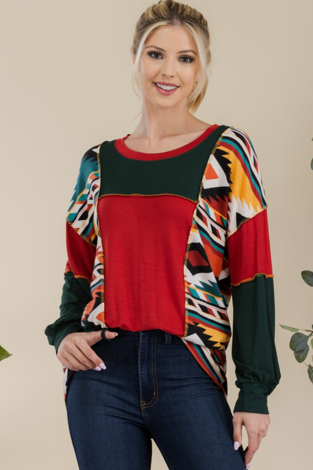 A woman stands against a plain background, wearing the Celeste Full Size Exposed Seam Printed Color Block T-Shirt with vibrant red, green, and yellow geometric sections, paired with dark jeans. She smiles, her hands resting in her jeans' pockets.
