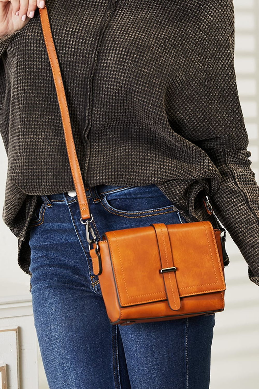 A person wearing a dark sweater and jeans holds the SHOMICO PU Leather Crossbody Bag, a stylish accessory in orange-brown featuring a flap and buckle detail.